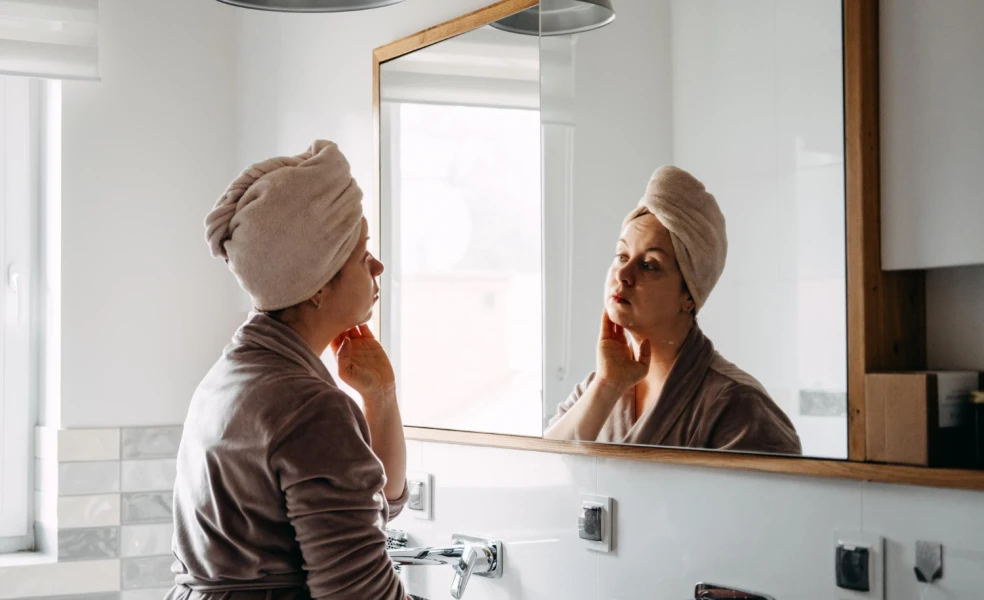 Žena v župane si pred zrkadlom kontroluje stav pleti počas skincare rutiny